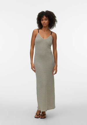 Görög hajú nő fekete-fehér, vízszintes csíkos, ujjatlan maxiruhát és fekete szandált visel, egyszínű háttér előtt áll.