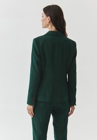 Zelený vlněný blazer s upraveným střihem, s klopami a zapínáním na knoflíky na rukávech. Textura je hladká, s jemnými švy na zadní straně.
