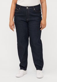 Plus-size person standing in dark denim jeans and white sneakers, wearing a light-colored top, with hands relaxed at their sides.