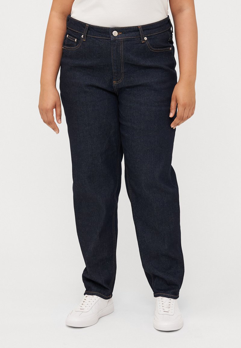 Plus-size person standing in dark denim jeans and white sneakers, wearing a light-colored top, with hands relaxed at their sides.