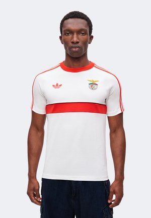 Joven con una camiseta blanca de Adidas con una franja horizontal roja, cuello rojo, rayas rojas en los hombros y un logo de un equipo deportivo en el pecho.