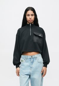 Γυναίκα με μακριά σκούρα μαλλιά, φορώντας ένα μαύρο κοντό φανελάκι με φερμουάρ και τσέπη, καθώς και ανοιχτό μπλε ψηλόμεσα τζιν, στέκεται μπροστά σε απλή φόντο.