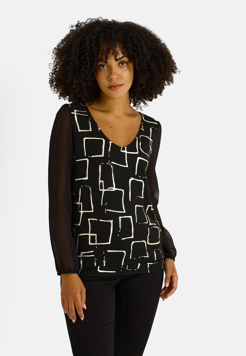 Blusa negra con patrones cuadráticos abstractos en blanco, escote en forma de V y mangas largas transparentes. La tela es suave con un ajuste ligeramente estructurado.