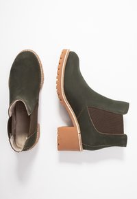 Botas de tobillo de cuero verde oscuro con un panel lateral elástico marrón, costuras beige y un tacón cuadrado marrón rugoso con surcos.