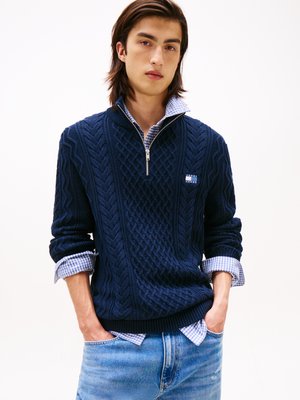 Pull en mailles torsadées bleu marine avec un col zippé, présentant un motif texturé et une petite étiquette. Porté par-dessus une chemise à carreaux bleu clair.