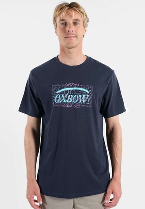 Homme blond portant un T-shirt bleu marine avec un logo "Oxbow Surfing Since 1985" en bleu clair et rose, debout devant un fond blanc uni.
