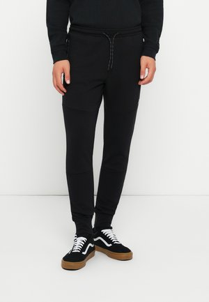 Zwarte sportbroek gemaakt van een gladde stof. Beschikt over een koord in de taille, zijzakken en een contrasterend paneelontwerp. Gepaard met zwarte sneakers.