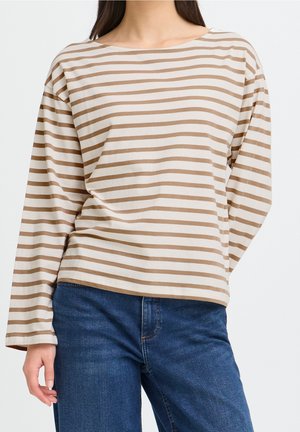 Femme portant un t-shirt à manches longues beige et blanc à rayures horizontales et un jean bleu, debout devant un fond uni.