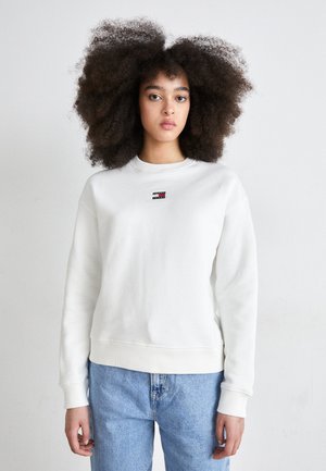 Felpa bianca con polsini e orlo a coste, caratterizzata da un piccolo logo multicolore sul petto. Abbinata a jeans di denim blu chiaro.