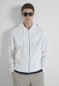 Una sudadera blanca con cremallera, hecha de tela suave, que cuenta con una capucha, dos bolsillos laterales y un logotipo en relieve en el pecho. Combinada con pantalones beiges.