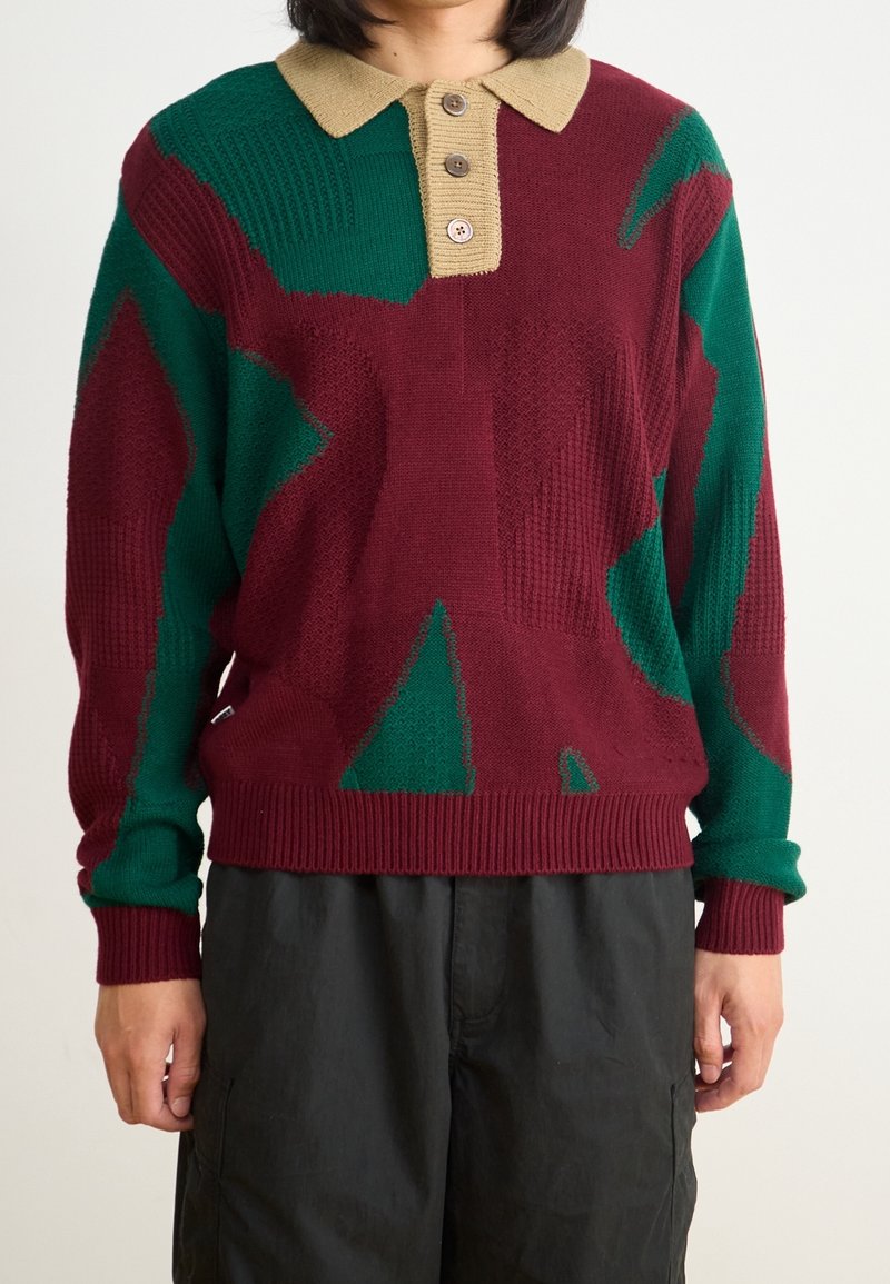 Personne portant un pull en tricot à motifs bordeaux et verts avec un col et des boutons beige, associé à un pantalon noir ample.