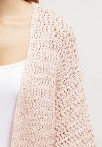 Zbliżenie na osobę noszącą jasnoróżowy kardigan z otwartym splotem na białym podkoszulku, pokazujące szczegółową teksturę splotu i obszar ramion.