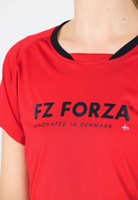 Röd sportt-shirt med svart krage, prydd med "FZ FORZA"-logotyp och texten "INNOVATED IN DENMARK" i svart. Slät tygstruktur.