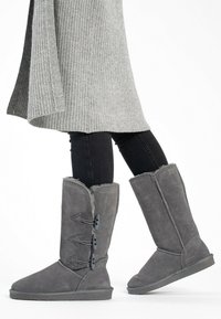 Stivali grigi in suede con fodera in pelliccia, altezza a metà polpaccio, caratterizzati da tre bottoni decorativi e una suola piatta. Indossati con jeans neri e una maglietta grigia.