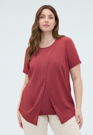 Kobieta długimi włosami, nosząca kolczyki-koła i warstwową bordową bluzkę z krótkim rękawem oraz beżowe spodnie, stojąca na jasnym tle.