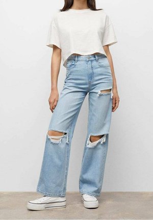 Beskåret beige t-shirt parret med højtrækkende lyseblå nedslidte jeans med store knærevner og frynsede kanter; hvide sneakers nedenunder.