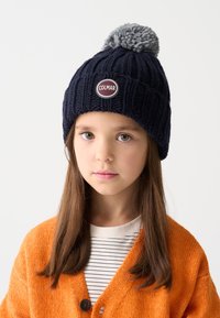 Caciula tricotată de culoare bleumarin cu un pompon gri deschis în vârf, decorată cu un patch metalic rotund inscripționat "COLMAR." Design texturat cu model de cablu.