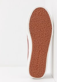 Sole of a sneaker with textured brown rubber grip and white edge, resting vertically against a white wall and surface.