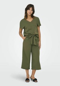 Conjunto de dos piezas en verde oliva con mangas cortas y cintura con lazo. Pantalones de pierna ancha, tejido suave y sandalias de tacón negro con detalles florales.