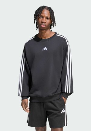 Joven con rastas cortas vistiendo sudadera negra de Adidas y pantalones cortos con rayas blancas sobre fondo liso.