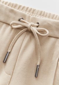Gros plan de la ceinture d'un pantalon de jogging beige avec un cordon noué et des embouts métalliques passant à travers des œillets argentés.