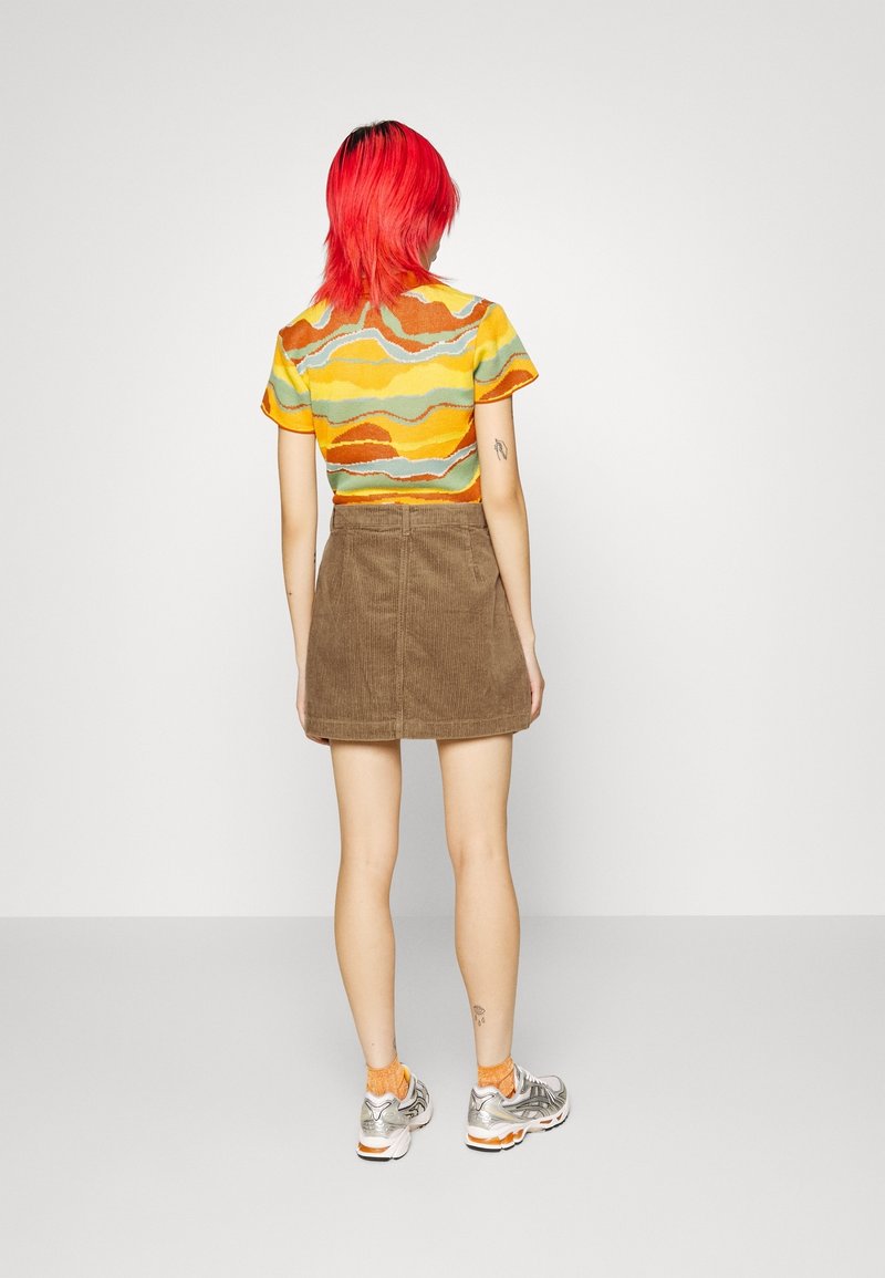 Stickad topp med vågig flerfärgad mönstring i gult, orange och grönt, kombinerad med en brun kordflamskjol och silverfärgade sportskor.