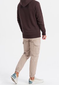 Brązowa bluza z kapturem, beżowe spodnie typu cargo z elastycznymi mankietami oraz białe trampki z niebieskimi i beżowymi akcentami. Gładkie tło.