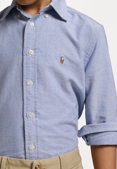 Ανοιχτό μπλε πουκάμισο με γιακά, φτιαγμένο από υφαντό ύφασμα, με λευκά κουμπιά και κεντημένο λογότυπο στο αριστερό στήθος. Κυρτωμένα μανίκια.