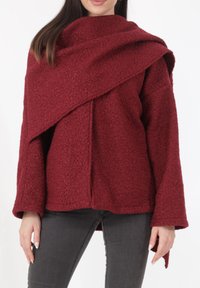 Bordeauxrode textuurlas met een asymmetrisch ontwerp, wijde mouwen en een gedrapeerde kraag, gecombineerd met nauwsluitende donkere spijkerbroek.