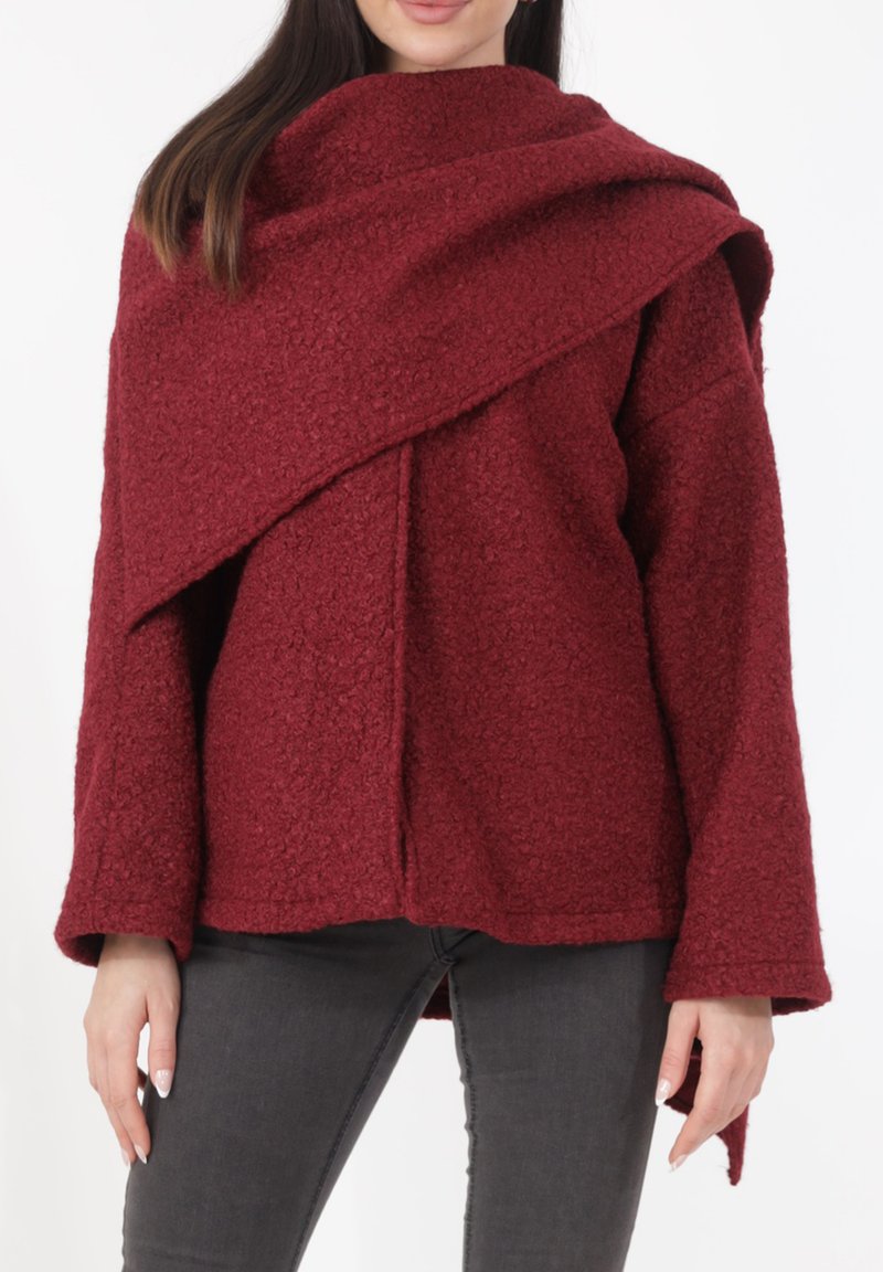 Bordeauxrode textuurlas met een asymmetrisch ontwerp, wijde mouwen en een gedrapeerde kraag, gecombineerd met nauwsluitende donkere spijkerbroek.