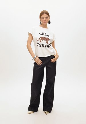 Camiseta blanca de manga corta con letras negras y un diseño de gato con estampado de leopardo. Combinada con pantalones anchos negros y zapatos metálicos.