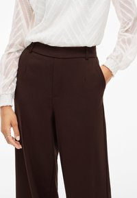 Ruske muško krojene hlače napravljene od glatke tkanine, s visokim strukom, bočnim džepovima i opuštenim krojem, uparenim s teksturiranom bijelom bluzom.