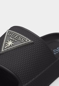 Εγγύς λήψη μαύρης διακοσμημένης σαγιονάρας με ασημένιο τρίγωνο λογότυπο "GUESS Beach" και έμβλημα φοίνικα στο λουρί.