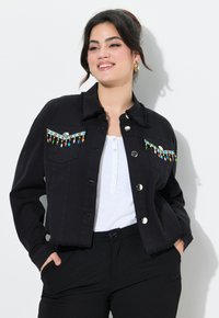 Musta lyhyt farkkutakki, jossa on fringed-yksityiskohtia ja värikkäitä helmikoristeita etutaskuissa, sisältäen hopeanväriset napit ja kauluksen.