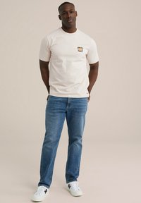 Heledad beež T-särk, millel on esikoti graafika, koos siniste teksapükste ja valgete tennisetossudega. Lihtne, läbilöön kangas lühikeste varrukatega.