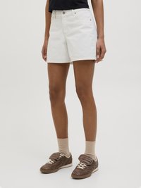 Λευκά τζιν σορτς με χαλαρή εφαρμογή, κλασικό σχεδιασμό πέντε τσεπών και κλείσιμο με κουμπί, σε συνδυασμό με καφέ αθλητικά παπούτσια και μπεζ κάλτσες.
