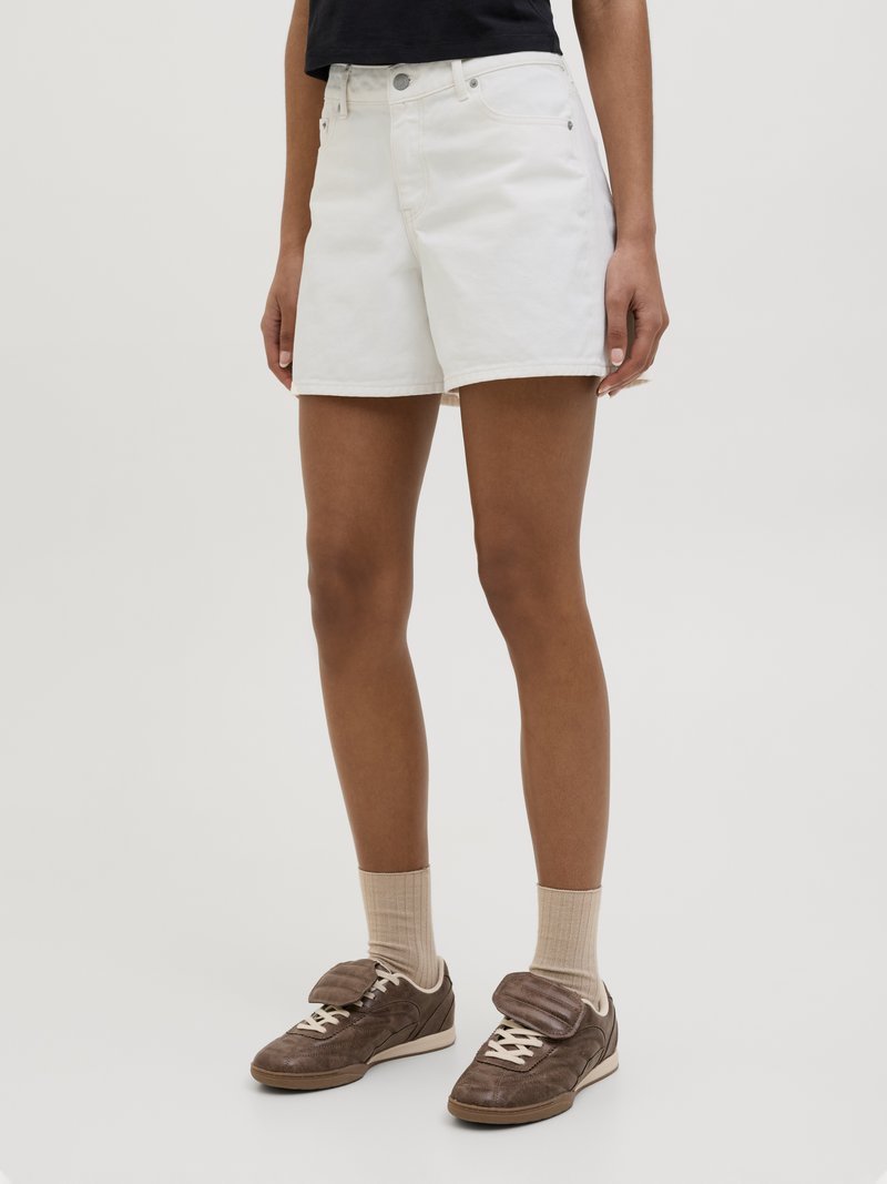 Λευκά τζιν σορτς με χαλαρή εφαρμογή, κλασικό σχεδιασμό πέντε τσεπών και κλείσιμο με κουμπί, σε συνδυασμό με καφέ αθλητικά παπούτσια και μπεζ κάλτσες.