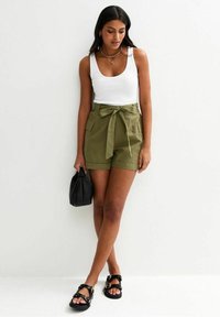 Olive green shorts with a bow tie at the waist, two front pockets, paired with a white tank top and black sandals, holding a black bag.