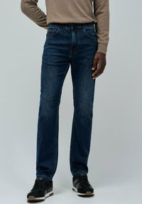 Vaqueros de mezclilla con un lavado azul oscuro, que cuentan con una cintura con cordón, cinco bolsillos y un corte recto, combinados con zapatillas negras.