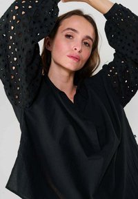 Eine Frau mit heller Haut und braunen Haaren trägt eine schwarze Bluse mit Puffärmeln im Lochmuster und posiert mit erhobenen Armen vor einem schlichten Hintergrund.
