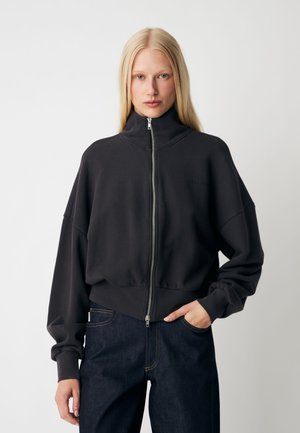 Vrouw met lang blond haar, gekleed in een zwarte jas met rits en donkerblauwe jeans, staat met één hand in de zak tegen een effen achtergrond.