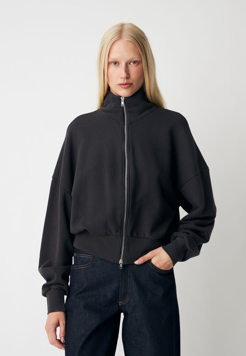 Femme aux longs cheveux blonds portant une veste noire zippée et un jean bleu foncé, debout avec une main dans la poche sur un fond uni.