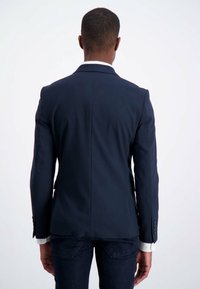 Uomo che indossa un blazer blu navy e jeans scuri, in piedi di spalle contro uno sfondo bianco uniforme.