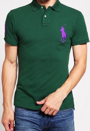 Homme portant un polo vert foncé bien ajusté avec un grand logo violet de joueur de polo sur la poitrine et un jean bleu, main reposant près de la taille.