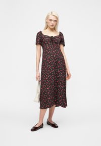 Νεαρή γυναίκα στέκεται φορώντας μαύρο μίντι φόρεμα με κόκκινο λουλουδάτο σχέδιο, φουσκωτά μανίκια και τετράγωνο ντεκολτέ, κρατώντας λευκή τσάντα χειρός και φορώντας μαύρες μπαλαρίνες.