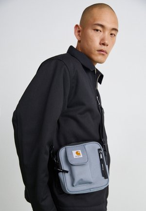 Homme à la tête rasée portant une veste noire et un sac bandoulière gris Carhartt sur un fond clair uni.