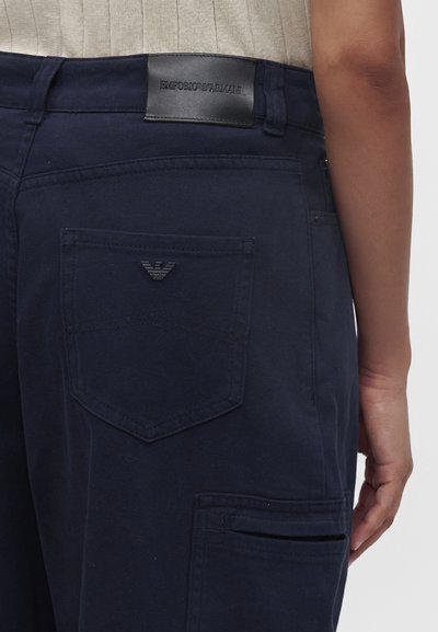 Άτομο που φορά ναυτικό μπλε παντελόνι Emporio Armani με ορατό λογότυπο στην πίσω τσέπη και μαύρη δερμάτινη ετικέτα επάνω από τη μέση.