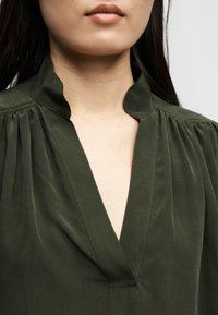 Γυναίκα που φοράει σκούρο πράσινο μπλουζάκι με V-λαιμόκοψη και μικρό όρθιο γιακά, που δείχνει τον λαιμό και το επάνω μέρος του στήθους της.