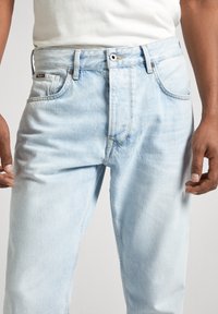 Hombre con vaqueros de denim azul claro y una camiseta blanca, con las manos relajadas a los lados, mostrado desde la cintura hasta la mitad del muslo.