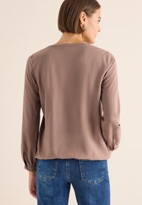 Blusa marrone a maniche lunghe realizzata in tessuto liscio, con scollo rotondo e polsini arricciati, abbinata a jeans in denim blu.